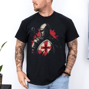 May include: A black t-shirt featuring a graphic of a knight in armour holding a shield with a red cross. The knight is wearing a helmet and has a sword in his hand.