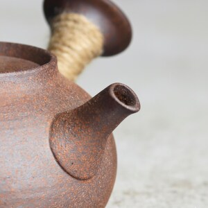 May include: Close-up of a brown ceramic teapot with a spout and a handle. The teapot has a textured surface and a rustic appearance. The handle is made of wood and wrapped with a natural fiber rope. The teapot is designed for brewing tea.