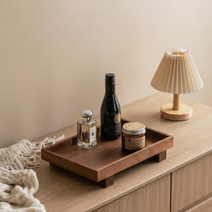 May include: A wooden tray with a dark finish holds a perfume bottle, a dark bottle, and a jar with a lid. A small lamp with a pleated shade sits on a wooden surface. The items are arranged on a wooden dresser.