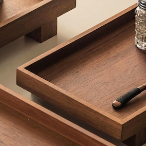 May include: Close-up of three wooden trays with a rich, dark brown finish. The trays have a rectangular shape with raised edges. One tray has a small wooden handle. The wood grain is visible, adding texture to the surface.