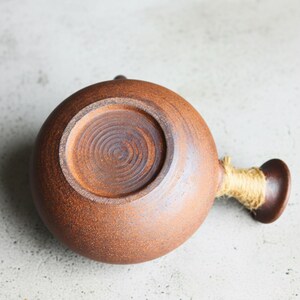 May include: A round, brown ceramic flask with a small handle and a spout wrapped in twine. The flask has a textured surface and a circular base with concentric grooves. The overall design is rustic and earthy.