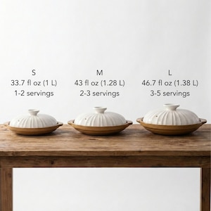 Op de afbeelding: Drie keramische kookpotten met deksels, in verschillende maten, worden getoond op een houten tafel. De potten zijn wit met geribbelde deksels en bruine bodems. Tekst boven de potten geeft maten aan: S (1 L), M (1,28 L) en L (1,38 L), met portie suggesties.