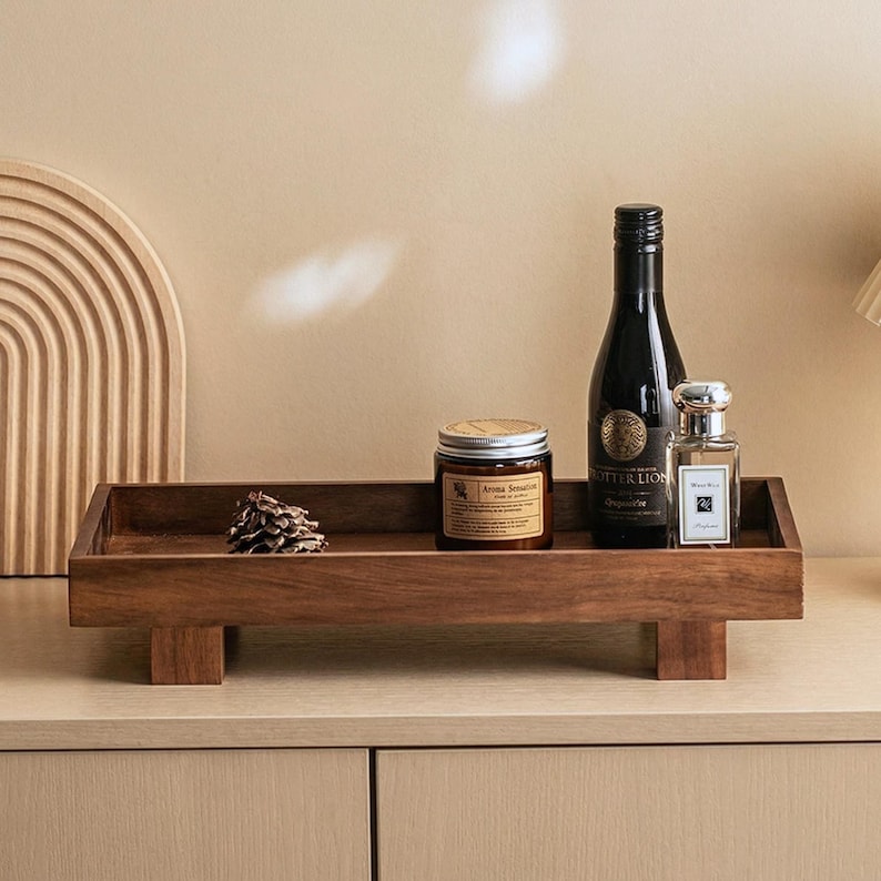May include: A rectangular, dark wood serving tray with a low profile, supported by four short legs. The tray holds a candle in a jar, a wine bottle, a perfume bottle, and a pine cone. The candle jar has the text "Aroma Sensation".