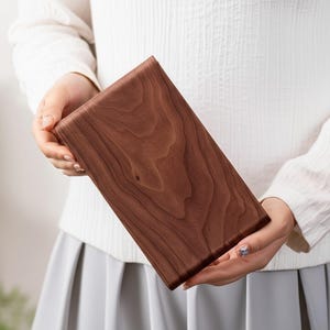 Peut inclure: Planche &agrave; d&eacute;couper rectangulaire en bois brun fonc&eacute;, avec des motifs de grain de bois visibles. La planche est tenue par deux mains, mettant en valeur sa surface lisse et polie. La planche mesure environ 30 cm de long et 20 cm de large.