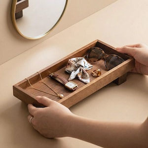 May include: A rectangular wooden tray holding accessories. The tray contains a gold necklace with a pearl, a brown leather watch, a patterned scarf, sunglasses, and a gold ring. The tray is being held by two hands.