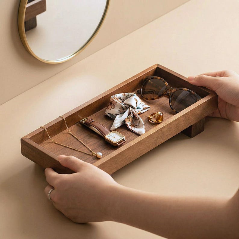 May include: A rectangular wooden tray holding accessories. The tray contains a gold necklace with a pearl, a brown leather watch, a patterned scarf, sunglasses, and a gold ring. The tray is being held by two hands.