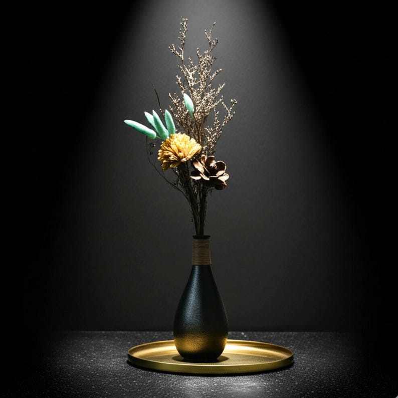Op de afbeelding: Een zwart-gouden vaas met gedroogde bloemen en bladeren, waaronder een gele bloem en een bruine dennenappel. De vaas staat op een goudkleurig dienblad, met een donkere achtergrond.