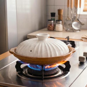 Op de afbeelding: Een witte keramische kookpot met deksel, op een gasfornuis met een blauwe vlam. De pot heeft een bruine rand en handvatten. Keukengerei en potten zijn zichtbaar op de achtergrond.