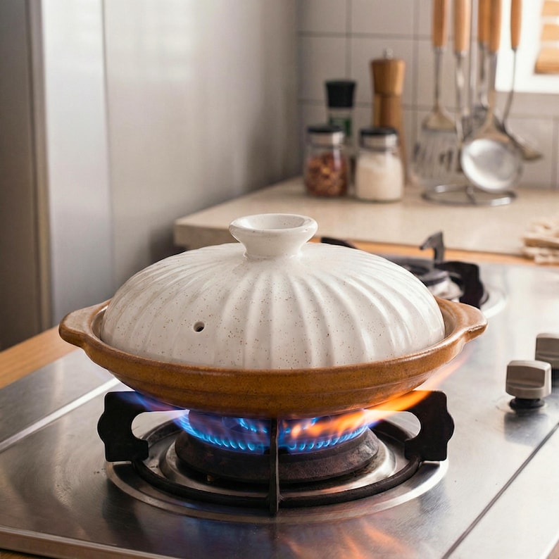 Op de afbeelding: Een witte keramische kookpot met deksel, op een gasfornuis met een blauwe vlam. De pot heeft een bruine rand en handvatten. Keukengerei en potten zijn zichtbaar op de achtergrond.