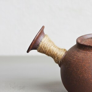May include: A brown ceramic teapot with a spout wrapped in natural twine. The teapot has a rounded body and a lid. The handle is a dark brown color, and the twine is a light tan color.