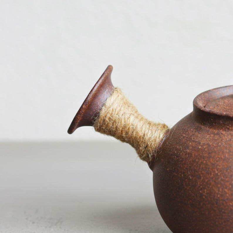 May include: A brown ceramic teapot with a spout wrapped in natural twine. The teapot has a rounded body and a lid. The handle is a dark brown color, and the twine is a light tan color.
