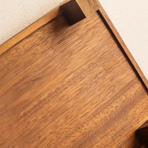 May include: Close-up of a wooden tray with a rich, warm brown color. The tray has a rectangular shape with a raised edge and a small, square wooden block at the corner. The wood grain is visible.