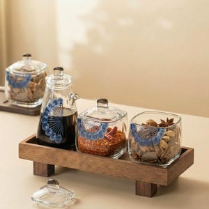 May include: A set of glass spice jars and a cruet with blue fan-shaped designs, filled with spices and liquids, on a wooden tray. The set includes a cruet with a spout and square jars with lids.