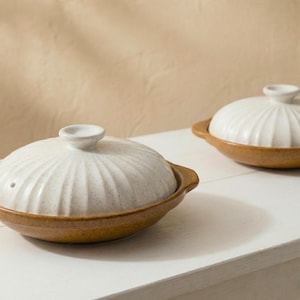 Op de afbeelding: Twee keramische kookpotten met deksels. De potten hebben een lichtbruine basis en een witte, geribbelde deksel met een klein handvat. De potten staan op een wit oppervlak, met een neutrale achtergrond.