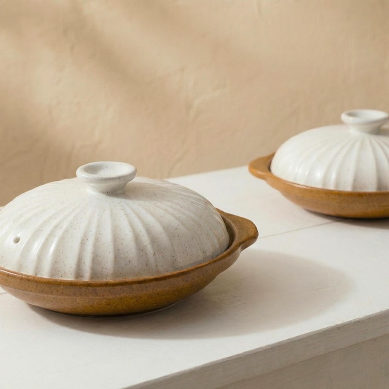 Op de afbeelding: Twee keramische kookpotten met deksels. De potten hebben een lichtbruine basis en een witte, geribbelde deksel met een klein handvat. De potten staan op een wit oppervlak, met een neutrale achtergrond.