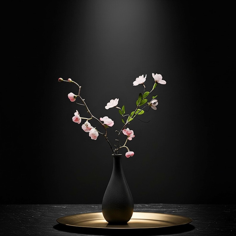 Op de afbeelding: Een zwarte vaas met een gouden voetstuk bevat een bloemstuk van roze bloesems en groene bladeren. De vaas staat op een goudkleurige schaal, alles tegen een donkere achtergrond. Het arrangement is een decoratief accent.