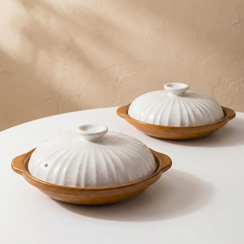 Op de afbeelding: Twee keramische kommen met deksel. De kommen hebben een lichtbruine basis en een witte, geribbelde deksel met een kleine knop. De kommen staan op een wit oppervlak, met een neutrale achtergrond.