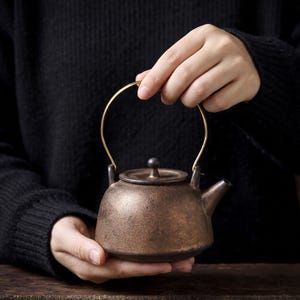 May include: A round, dark bronze-colored ceramic teapot with a textured, weathered surface. It features a small, matching lid with a central knob, a short spout, and a thin, arched gold-toned metal handle attached to two side loops.