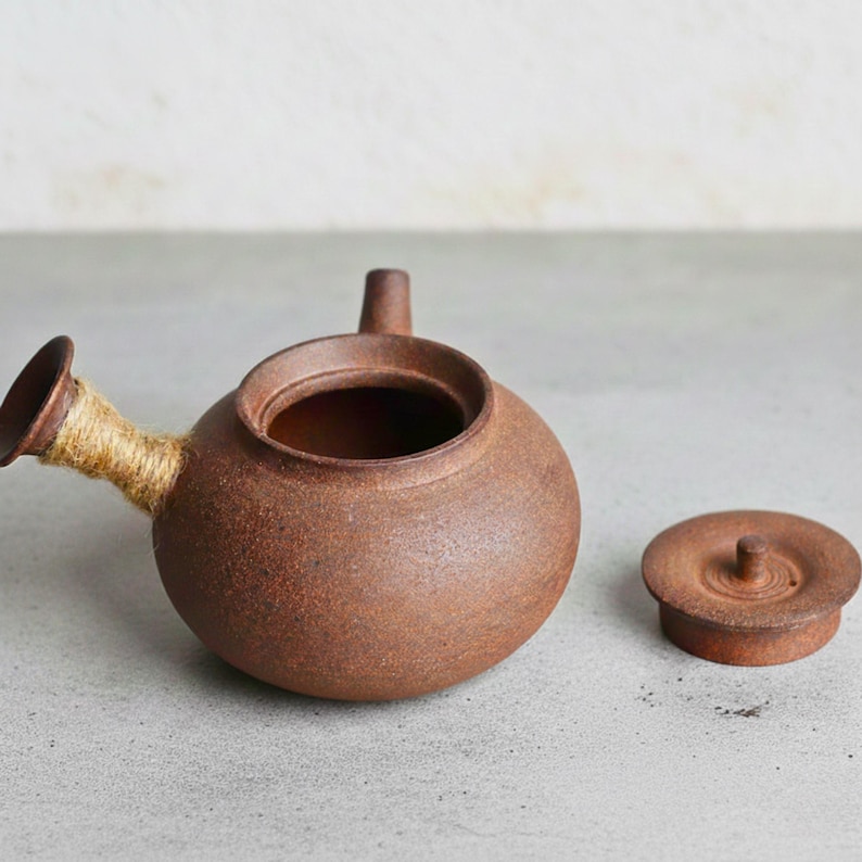 May include: A small, round, brown ceramic teapot with a spout wrapped in tan twine and a separate lid. The teapot has a short handle and a small, cylindrical spout. The lid is flat with a small knob.