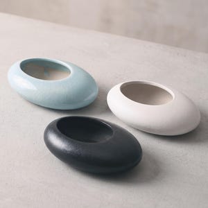 Puede incluir: Tres macetas de cerámica de forma ovalada con bordes suaves y redondeados. Una maceta presenta un acabado azul claro brillante con un patrón de grietas finas, otra un acabado blanco mate y la tercera un acabado negro carbón texturizado.