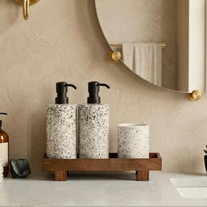 May include: Bathroom accessories set featuring two speckled soap dispensers with black pumps, a matching tumbler, and a wooden tray. The set is designed with a modern aesthetic, perfect for a minimalist bathroom decor.