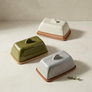 May include: Three ceramic butter dishes in olive green, speckled gray, and off-white. Each dish has a rectangular base and a domed lid with a small handle. The bases are a natural terracotta color.