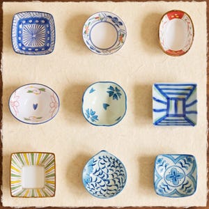 Puede incluir: Nueve pequeños platos de cerámica decorativos con varios patrones y formas. Los platos presentan diseños azules, blancos, verdes y amarillos, incluyendo motivos florales, geométricos y abstractos. Están dispuestos sobre una superficie clara y texturizada.