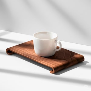 Op de afbeelding: Een witte keramische mok met een gebogen handvat staat op een rechthoekige, donkerbruine houten dienblad. De lade heeft aan elk uiteinde een gebogen, verhoogde rand. De mok en de lade staan tegen een witte achtergrond met schaduwen.
