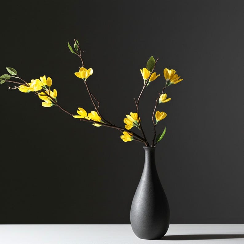 Op de afbeelding: Een zwarte keramische vaas bevat takken met heldergele bloemen en groene bladeren. De vaas heeft een slanke hals en een afgeronde basis. De achtergrond is donkergrijs, wat een contrast cre&euml;ert met het bloemstuk.