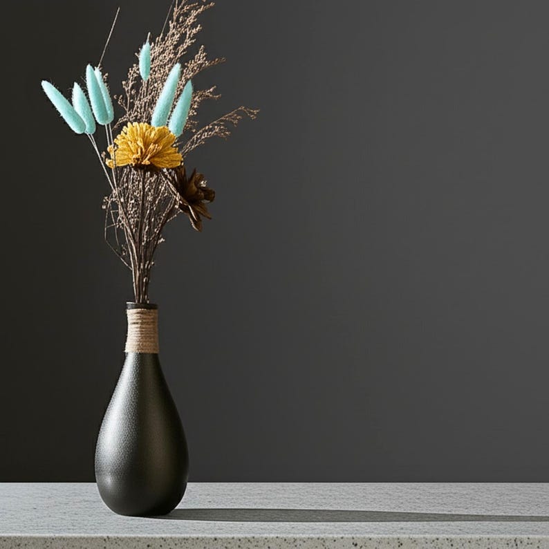 Op de afbeelding: Een zwarte, druppelvormige vaas met een getextureerd oppervlak, omwikkeld met touw bij de hals, bevat een boeket droogbloemen. Het arrangement bevat gele, blauwe en bruine elementen, tegen een donkergrijze achtergrond.
