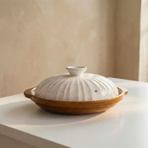 May include: A white ceramic lidded pot with a brown base sits on a white surface. The lid has a ribbed design and a small handle. The pot is likely used for cooking and serving food.