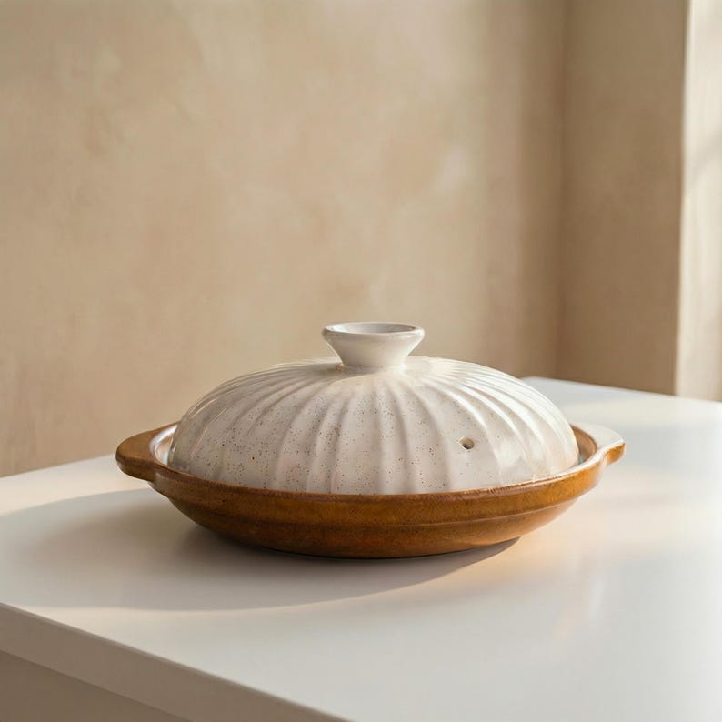 Op de afbeelding: Een witte keramische pot met deksel en bruine basis staat op een wit oppervlak. Het deksel heeft een geribbeld ontwerp en een kleine handgreep. De pot wordt waarschijnlijk gebruikt om eten in te koken en te serveren.