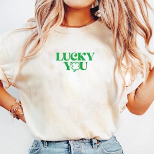 Può includere: Una maglietta bianca con il testo "LUCKY YOU" in lettere verdi e un quadrifoglio verde.