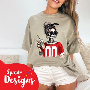 Peut inclure: Une femme portant un t-shirt gris avec un motif de squelette portant un maillot rouge et tenant une tasse de caf&eacute;. Le motif de squelette a un bandana noir et des lunettes de soleil.