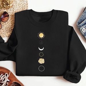 May include: Black sweatshirt featuring a gold sun, moon, and flower design. The design is vertically aligned in the centre of the sweatshirt.