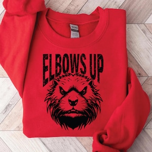Puede incluir: Sudadera roja con un gráfico negro de un castor con el texto "Elbows Up" impreso en ella.