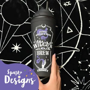 Puede incluir: Taza de viaje reutilizable negra con un dise&ntilde;o blanco que dice "Freshly Bottled & Hexed Witch's Brew" con una luna creciente morada, estrellas y una mano sosteniendo una poci&oacute;n. La taza tiene una tapa negra y una pajita transparente.