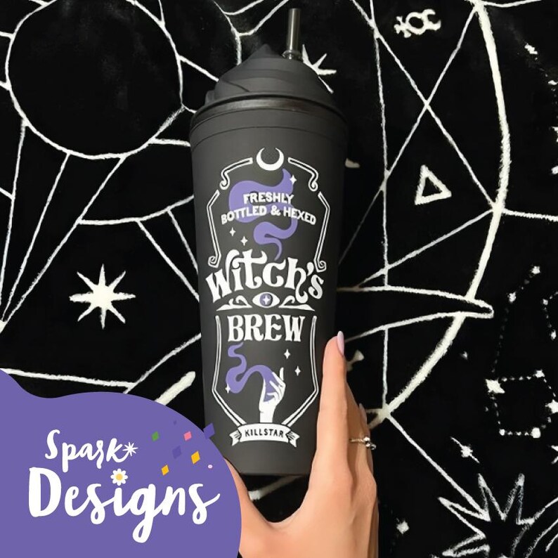 Puede incluir: Taza de viaje reutilizable negra con un dise&ntilde;o blanco que dice "Freshly Bottled & Hexed Witch's Brew" con una luna creciente morada, estrellas y una mano sosteniendo una poci&oacute;n. La taza tiene una tapa negra y una pajita transparente.