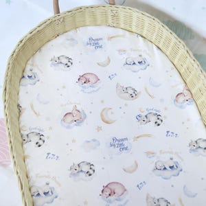 May include: A white wicker bassinet with a white and blue patterned liner. The liner features a repeating pattern of sleeping animals, stars, and moons. The text "Dream big little one" is repeated on the liner.
