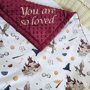 May include: A burgundy baby blanket with the embroidered phrase "You are so loved." The blanket features a pattern of owls, eyeglasses, wands, and a castle on a light gray background. The blanket is folded over a white surface.