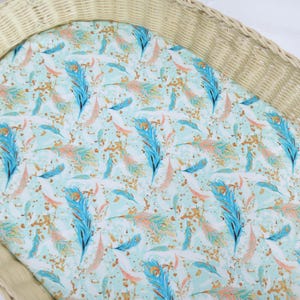 May include: A white oval-shaped wicker basket with a fitted sheet featuring a turquoise and gold feather pattern.