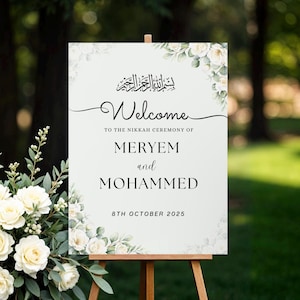 Può includere: Un cartello di benvenuto per matrimoni bianco su un cavalletto di legno. Il cartello presenta accenti floreali ed un'elegante calligrafia che recita "Benvenuti" e i nomi "MERYEM e MOHAMMED" con la data "8 ottobre 2025".