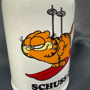 May include: White ceramic mug featuring Garfield the cat skiing on a red ski. The orange cat is wearing a ski outfit and skis. The word "SCHUSS" is printed in black below the cat.