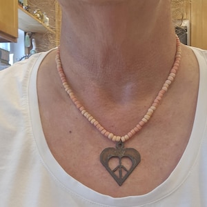 May include: A necklace featuring a series of small, round, light peach-colored beads strung together. The necklace includes a dark, metallic, heart-shaped pendant with a cutout peace sign symbol centered within the heart frame.
