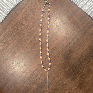 May include: A beaded necklace featuring alternating peach and periwinkle blue cylindrical beads strung on a cord. The necklace includes a silver-toned metal toggle clasp at the top and a long, slender, pointed silver-toned metal pendant at the bottom.