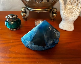 May include: A large, faceted blue crystal with a rough, natural base sits on a wooden surface. The crystal's color transitions from a deep blue to a lighter shade. Other decorative items are in the background.