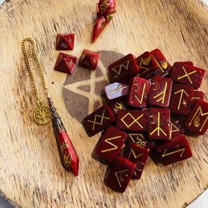 May include: A collection of divination tools, including red resin runes with gold symbols, a pendulum with a gold chain and tree of life charm, and a red bird skull. The items are arranged on a wooden tray.