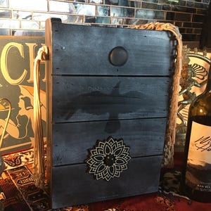 May include: A black wooden box with a rope handle and a black mandala design. The box has a black bird silhouette and a black button on the front.