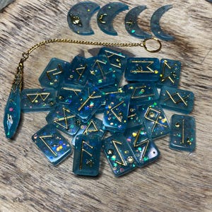 May include: A set of blue and gold rune stones with a gold chain and three crescent moon charms. The runes are set on a wooden surface.