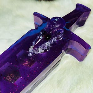 May include: A purple resin dagger with embedded dice and glitter. The dagger has a crossguard and a rounded pommel. The resin is a deep purple with blue accents and is encrusted with small crystals. The dice are visible within the resin.